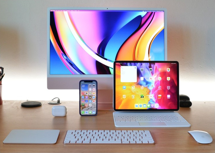 Apple desktop, laptop, and iPhone on wooden desk.