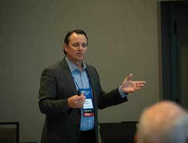 A man in a suit presents in a conference room, gesturing with one hand.