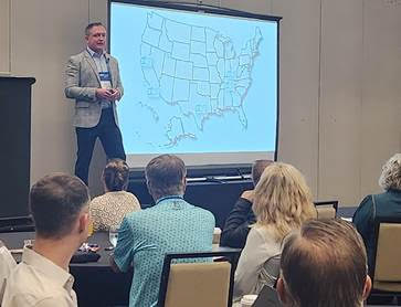 A man in a suit presents in front of a projected U.S. map to a seated audience in a conference room.