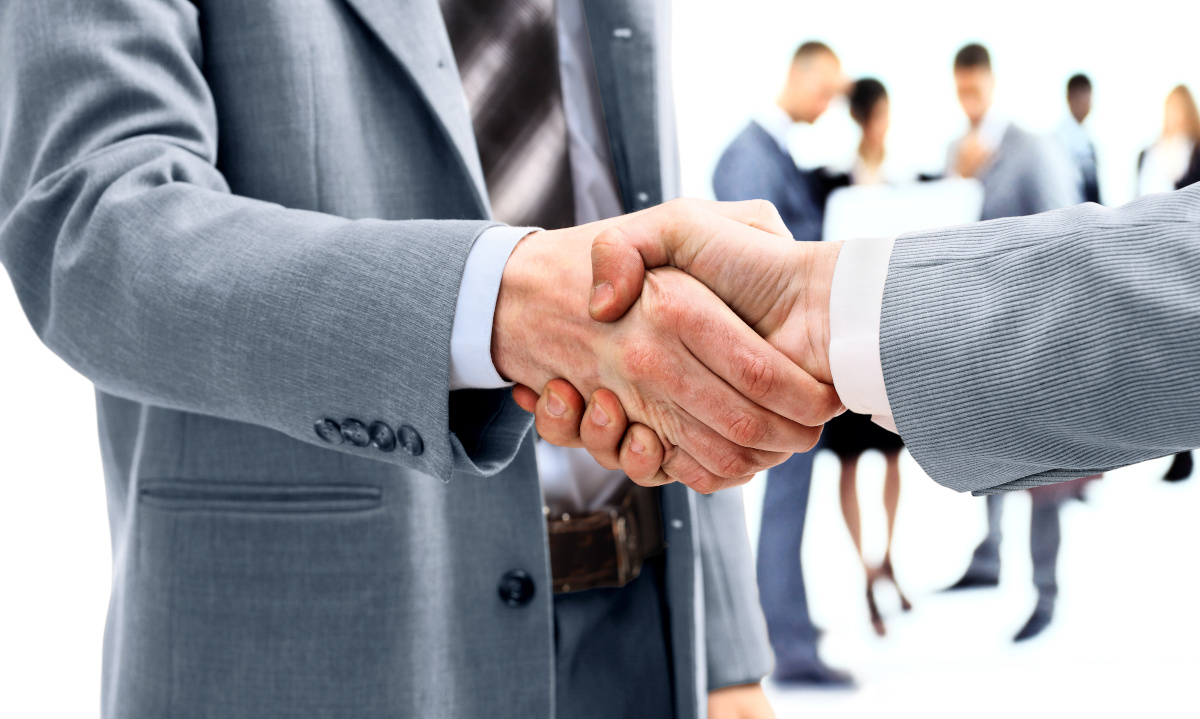 A close up on two men in suits shaking hands, with a group of people working blurred in the background