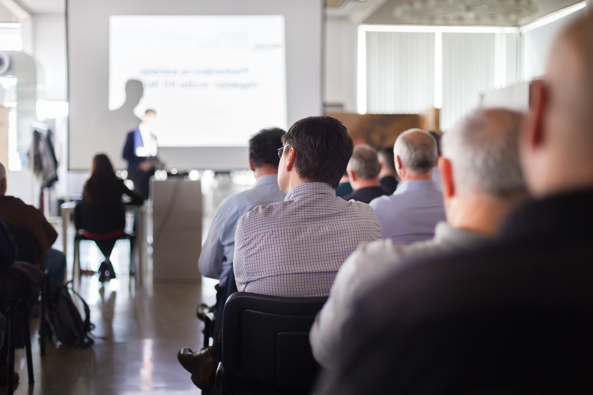 Audience at a seminar hall listening to a presentation. The speaker stands at the front, and a bright screen displays blurred text. Professional atmosphere.